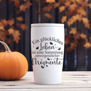 May include: A white travel mug with a lid, featuring black text that reads "Ein gl&uuml;ckliches Leben ist eine Sammlung unvergesslicher Momente" and butterfly illustrations. An orange pumpkin sits to the left of the mug. The background is blurred.