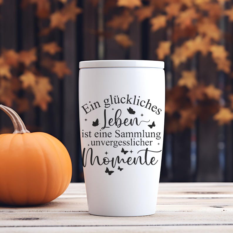 May include: A white travel mug with a lid, featuring black text that reads "Ein gl&uuml;ckliches Leben ist eine Sammlung unvergesslicher Momente" and butterfly illustrations. An orange pumpkin sits to the left of the mug. The background is blurred.