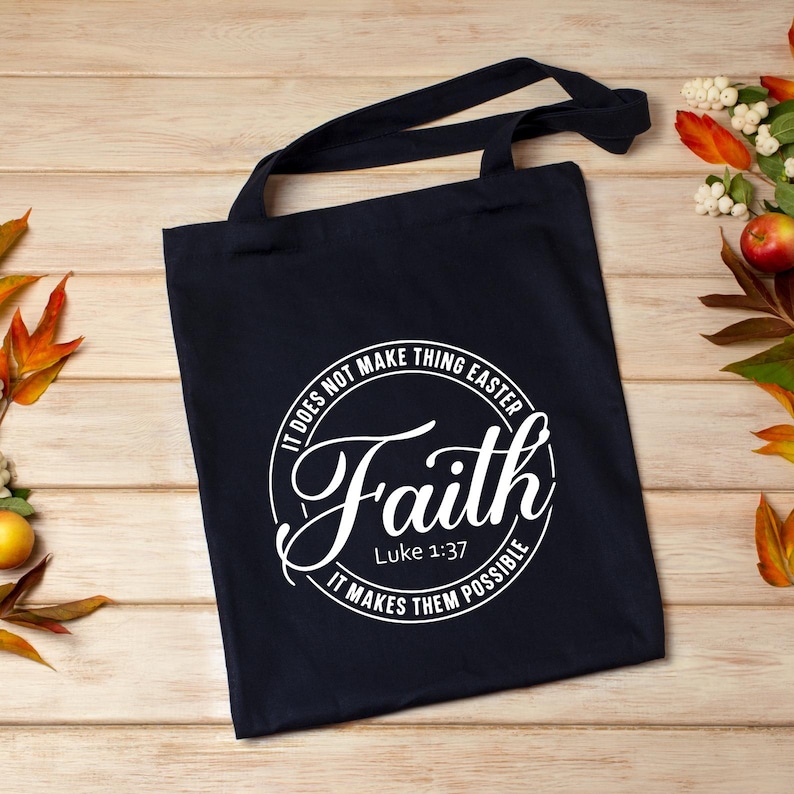 Peut inclure: Un sac fourre-tout bleu marine avec des poign&eacute;es noires. Le sac pr&eacute;sente un motif circulaire blanc avec le mot "Faith" en &eacute;criture cursive, et le texte "It does not make things easier, it makes them possible" et "Luke 1:37".