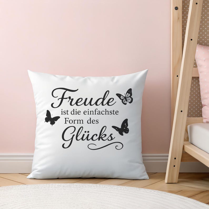 May include: White square pillow with black text and butterfly illustrations. The text reads "Freude ist die einfachste Form des Gl&uuml;cks". The pillow is on a light pink background.