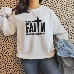 Peut inclure: Sweat-shirt blanc avec l'inscription noire "WALKING BY FAITH TRIPPING CONSTANTLY". Le motif comprend une croix. Le sweat-shirt a des manches longues et un col rond.