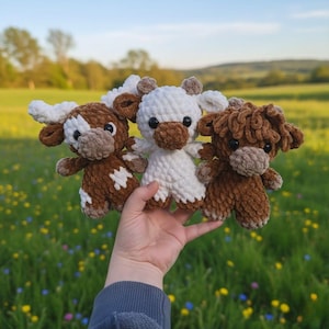 Peut inclure: Trois peluches de vaches au crochet faites à la main, marron, blanches et un mélange des deux. Les peluches ont de petites cornes, des visages bruns et sont tenues devant un arrière-plan flou d'un champ avec des fleurs jaunes et des arbres.
