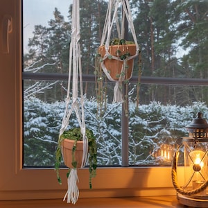 May include: Two terracotta pots with greenery hang from white macrame plant hangers in front of a window. The pots are adorned with green beaded strands. A lit lantern sits on the windowsill, and a snowy forest is visible outside.