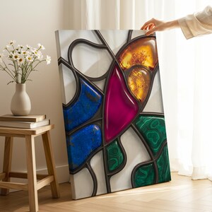May include: A canvas art piece featuring a stained glass design with vibrant colors. The artwork showcases sections of blue, magenta, green, and amber hues, outlined with dark gray lines. The piece is held by a person's hand.