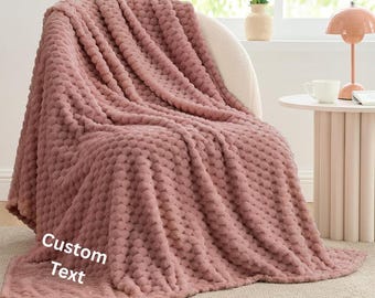 May include: A dusty rose-colored blanket with a textured, bubble-like pattern draped over a chair. The blanket has a soft, plush appearance. The words "Custom Text" are visible on the blanket.