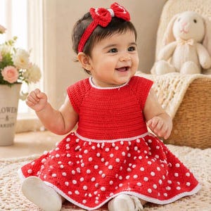Könnte beinhalten: Ein Baby trägt ein rotes gehäkeltes Kleid mit weißen Punkten und ein passendes Stirnband mit Schleife. Das Kleid hat einen weißen Saum und das Baby lächelt. Ein Stoffhase ist im Hintergrund.