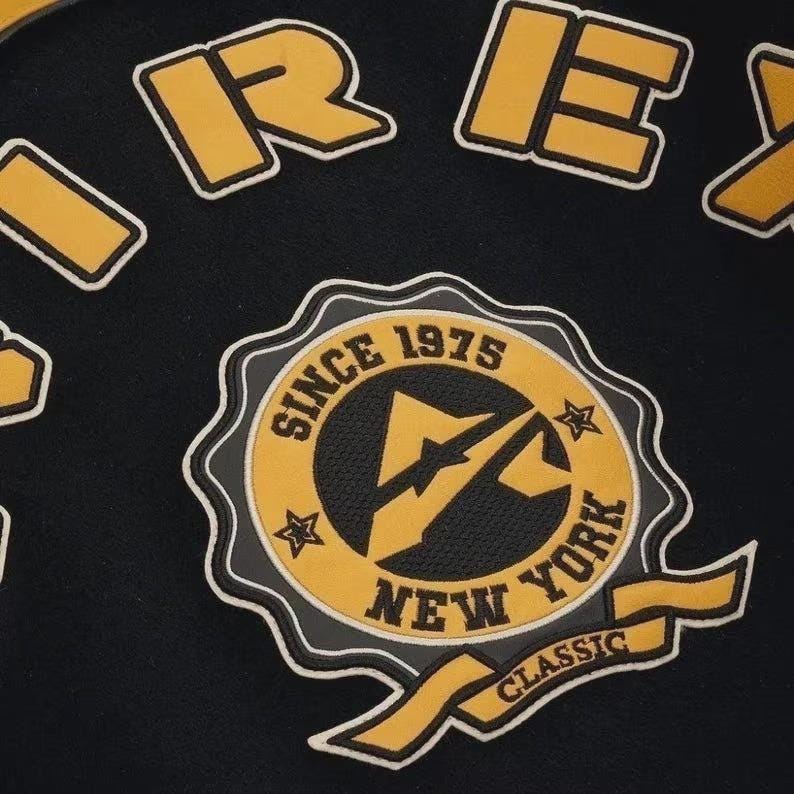 May include: Black fabric featuring yellow and white embroidered patches. The central patch reads "Since 1975 New York Classic" with a lightning bolt design. Other patches spell out "REX" in yellow block letters.