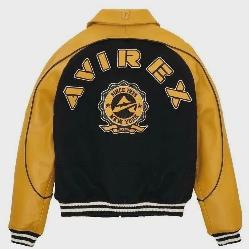 May include: Varsity jacket with a black back panel and mustard yellow sleeves and collar. The back features the word "AVIREX" in a varsity font above a circular emblem with the text "SINCE 1975 NEW YORK CLASSIC". The jacket has a striped hem and cuffs.