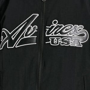 May include: Black jacket with white embroidered text that reads "X-iness USA". The jacket has a zipper closure and a sporty design. The text is in a stylized font.