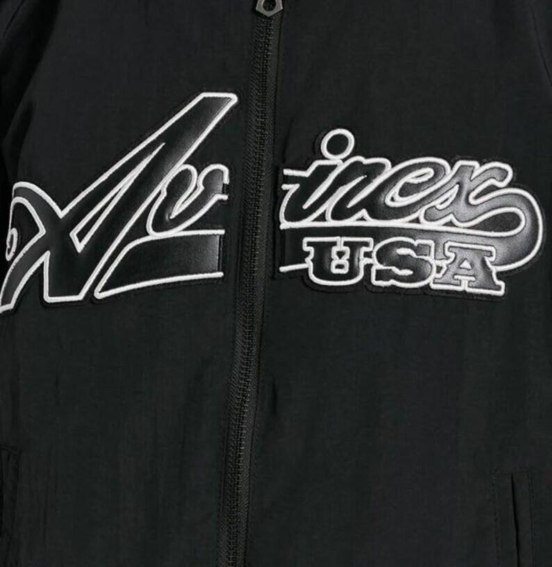 May include: Black jacket with white embroidered text that reads "X-iness USA". The jacket has a zipper closure and a sporty design. The text is in a stylized font.