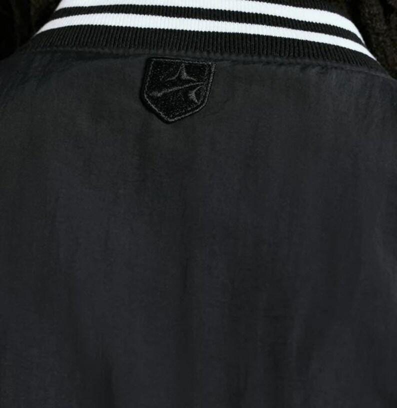 May include: Black bomber jacket with a white and black striped collar. The jacket features a small, black emblem on the upper back. The jacket is made of a smooth, possibly nylon, material.