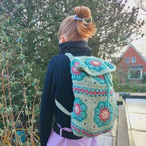 Könnte beinhalten: Handgefertigter Häkelrucksack in Grün-, Rosa- und Türkistönen mit floralem Muster. Der Rucksack hat einen Kordelzugverschluss und unifarbene grüne Träger. Der Rucksack wird über der Schulter getragen.