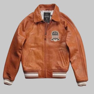 May include: A brown leather jacket with a textured finish, collared neckline and zip-up front. The jacket has a patch on the chest with the text "AVIREX" and a striped design on the cuffs and hem.