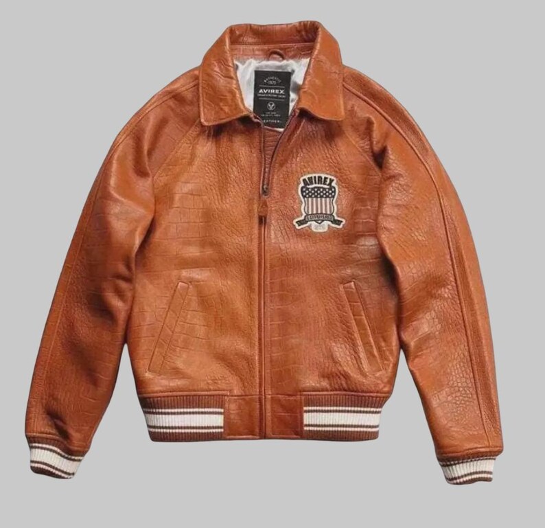 May include: A brown leather jacket with a textured finish, collared neckline and zip-up front. The jacket has a patch on the chest with the text "AVIREX" and a striped design on the cuffs and hem.