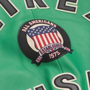May include: Close-up of a green jacket with a circular patch featuring the American flag and the text "ALL AMERICANS ESTABLISHED 1975 NY". Black and white lettering is visible on the jacket.