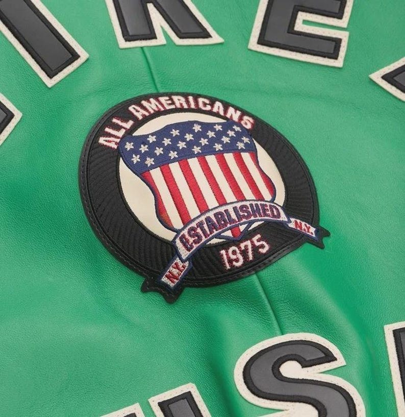 May include: Close-up of a green jacket with a circular patch featuring the American flag and the text "ALL AMERICANS ESTABLISHED 1975 NY". Black and white lettering is visible on the jacket.