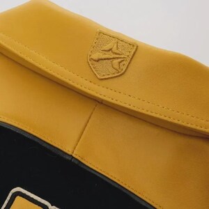 May include: Close-up of a mustard yellow leather item with a stitched emblem. The emblem is a shield shape with a detailed design. The item has a black section at the bottom.