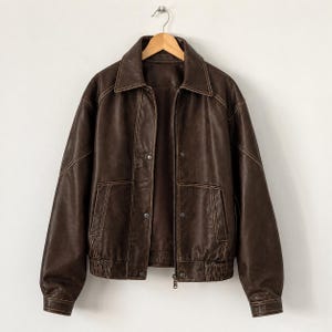 Könnte beinhalten: Braune Lederjacke mit Kragen, Knopfverschluss und zwei Vordertaschen. Die Jacke hat einen gerippten Saum und Bündchen und hängt an einem Holzbügel. Die Nähte sind hellbraun.