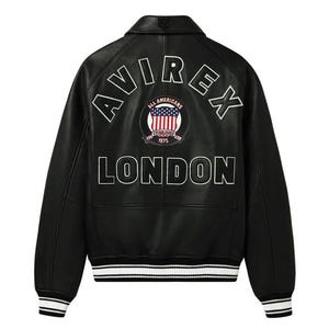 Könnte beinhalten: Schwarze Lederjacke mit weißem Schriftzug "AVIREX" und "LONDON". Ein kreisförmiger Aufnäher zeigt die amerikanische Flagge und die Worte "ALL AMERICANS ESTABLISHED 1975". Die Jacke hat einen weiß-schwarzen Streifen.