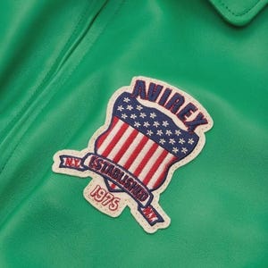 May include: Close-up of a green leather jacket with a patch. The patch features a shield design with red and white stripes, blue stars, and the text "AVIREX NY ESTABLISHED 1975". The jacket has a collar.