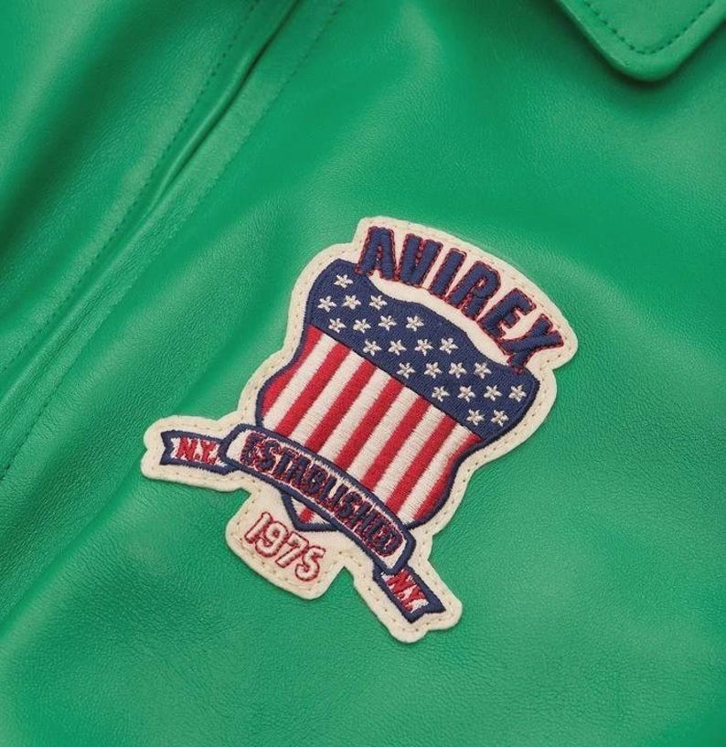 May include: Close-up of a green leather jacket with a patch. The patch features a shield design with red and white stripes, blue stars, and the text "AVIREX NY ESTABLISHED 1975". The jacket has a collar.