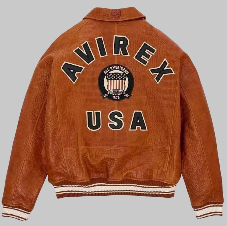 May include: A brown leather bomber jacket featuring the word "AVIREX" in black and white lettering across the back. Below is a patch with "ALL AMERICANS" and "ESTABLISHED 1975". The word "USA" is below the patch. The jacket has a ribbed collar and cuffs.