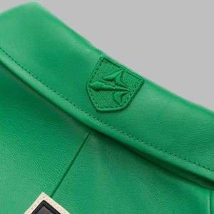 May include: Close-up of a bright green leather item with a stitched seam. A green embroidered emblem, possibly a logo, is sewn onto the item. The emblem is a shield shape with a stylized design.