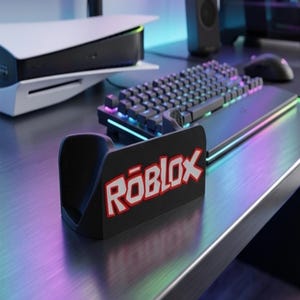 May include: A black stand with the Roblox logo in red and white is on a desk. A black keyboard with colorful lights is in the background, along with a white and black gaming console.