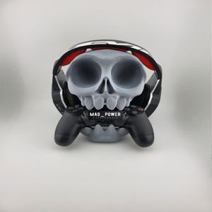 Puede incluir: Un soporte para auriculares con forma de calavera gris que sostiene un mando de juego negro. El soporte tiene cuencas de ojos grandes y una mandíbula detallada. Los auriculares son negros y blancos con detalles en rojo. El texto "MAD_POWER" es visible en el mando.