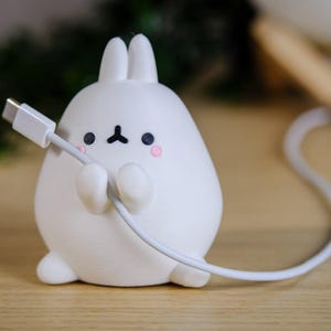 Könnte beinhalten: Ein weißer, hasenförmiger Kabelorganizer mit einem Ladekabel. Der Hase hat schwarze Augen, eine schwarze Nase und rosa Wangen. Das Kabel ist weiß und wird von den Armen des Hasen gehalten. Der Hase sitzt auf einer Holzoberfläche.