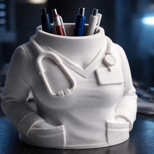 Pode incluir: Um porta-canetas branco em forma de jaleco médico, completo com estetoscópio e etiqueta de identificação. O porta-canetas está cheio de canetas de várias cores, incluindo vermelho, azul e preto. Um acessório de mesa único para profissionais de saúde.