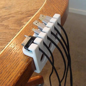 Pode incluir: Um organizador de cabos branco com cinco ranhuras, segurando vários cabos de carregamento USB e de dispositivos. O organizador está preso à borda de uma mesa de madeira. Cabos pretos se estendem do organizador.