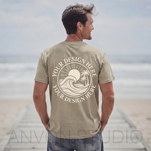 May include: A khaki-colored t-shirt with a circular graphic on the back featuring a sun, waves, and palm trees. The text "YOUR DESIGN HERE" is printed around the graphic. The shirt is being worn by a person on a beach.
