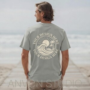 May include: A light green t-shirt with a circular design on the back featuring a sun, waves, and palm trees. The text "YOUR DESIGN HERE" is printed around the design. The shirt is being worn on a beach.