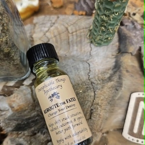 May include: A small glass bottle of ritual oil with a black cap and a beige label. The label reads Inexplicable Things Apothecary, REROUTE THE FATES, Change Your Destiny, Helps shift stuck situations, redirect outcomes, & open a better path forward, Apply with adaptability, 1/2 fl oz Ritual Oil.