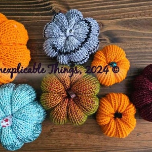 May include: Six knitted pumpkins in various colors, including orange, teal, burgundy, gray, and green. The pumpkins are arranged on a wooden surface. Inexplicable Things, 2024 is written on the surface.