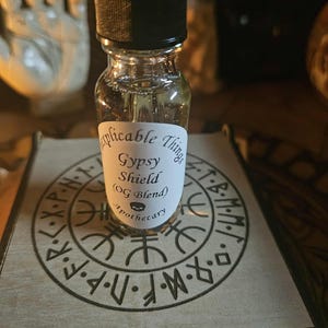 Peut inclure: Petite bouteille en verre avec un bouchon compte-gouttes noir, étiquetée "Applicable Things Gypsy Shield (OG Blend) Apothecary". La bouteille repose sur une boîte en bois avec un motif circulaire gravé. Le liquide à l'intérieur est jaune clair.