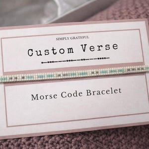 Puede incluir: Una pulsera con código Morse descansa sobre una tarjeta que dice "Custom Verse" y "Morse Code Bracelet". La pulsera está hecha de pequeñas cuentas de color claro con un patrón de cuentas azules y grises. La tarjeta es de color rosa claro con un borde simple.