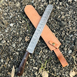 Peut inclure: Un long couteau en acier de Damas avec un manche en bois foncé et un étui en cuir beige. La lame présente un motif distinctif et l'étui est doté d'une fermeture à bouton. Le couteau est conçu pour couper.