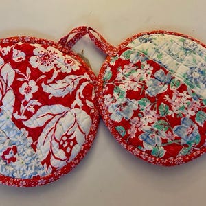 May include: A pair of round, quilted pot holders connected by a fabric loop. The fabric features a vibrant red background with white floral patterns and contrasting light blue and white sections. The edges are finished with a red floral trim.