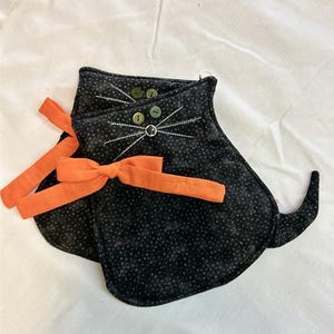 May include: Two black cat-shaped oven mitts with white stitched whiskers and button eyes. Each mitt has an orange bow. The mitts are made of black fabric with a white polka dot pattern.