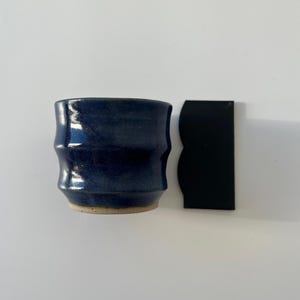 Op de afbeelding: Een handgemaakte, donkerblauwe keramische beker met een geribbeld ontwerp. De beker heeft een lichtbruine basis en een glanzende afwerking. Naast de beker ligt een zwart, rechthoekig object met een gebogen rand.