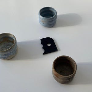 Peut inclure: Trois petits récipients en céramique bleus, bruns et beiges, ainsi qu'un outil noir de forme irrégulière avec un trou central. Les récipients ont un aspect texturé, suggérant une qualité artisanale. Les objets sont disposés sur une surface blanche.
