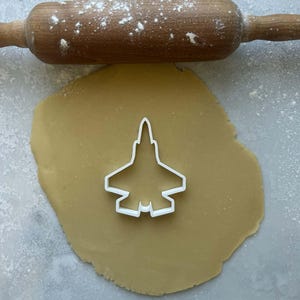 May include: A white plastic cookie cutter in the shape of a fighter jet sits on a sheet of dough. A wooden rolling pin dusted with flour rests above the dough. The dough is light brown.