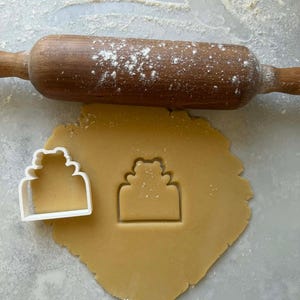 Peut inclure: Un rouleau à pâtisserie en bois saupoudré de farine repose au-dessus d'un biscuit découpé et d'un emporte-pièce en plastique blanc. L'emporte-pièce et le biscuit découpé ont la forme d'un gâteau à étages. La pâte est brun clair.