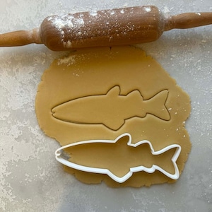 May include: A wooden rolling pin dusted with flour rests above a sheet of dough. A fish-shaped cookie cutter sits on the dough, next to an impression of the same shape. The dough is light brown.