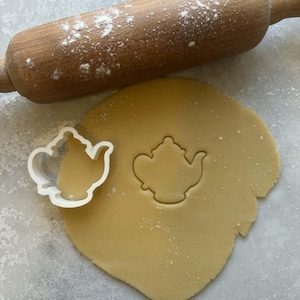 Può includere: Un mattarello di legno spolverato di farina è appoggiato su un foglio di pasta. Un tagliabiscotti bianco a forma di teiera in plastica è accanto a un'impronta della stessa forma nella pasta. Tema della panificazione e della cucina.