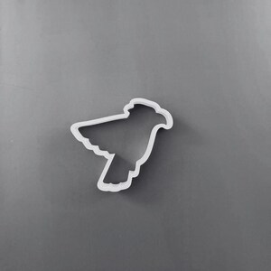 Puede incluir: Un cortador de galletas de plástico blanco con forma de pájaro en vuelo, con las alas extendidas hacia arriba y las plumas de la cola apuntando hacia abajo. El contorno del pájaro está claramente definido por los bordes elevados.