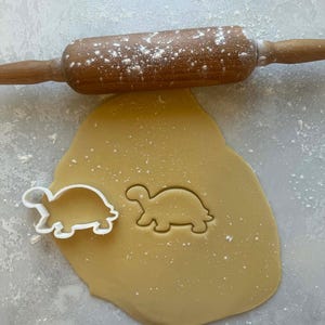 May include: A wooden rolling pin dusted with flour sits above a sheet of dough. A turtle-shaped cookie cutter sits next to a turtle-shaped impression in the dough. The dough is light brown, and the rolling pin is a medium brown.