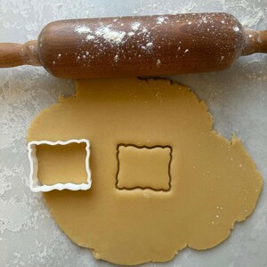 Puede incluir: Un rodillo de madera espolvoreado con harina descansa sobre una lámina de masa. Un cortador de galletas rectangular blanco con un borde ondulado se encuentra junto a una forma recortada en la masa. Concepto de repostería.